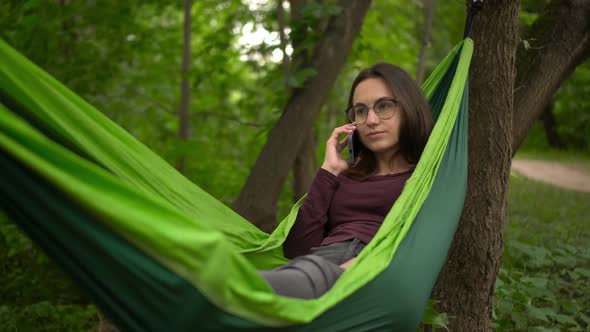 A Young Woman Swings in a Hammock and Talks on the Phone alt
