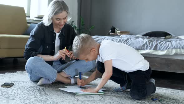 A Young Mother and a Baby Boy Are sitting on the Floor, The Boy Is Drawing with Felt-Tip Pens alt