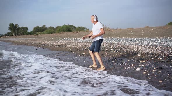 A Slender Whitehaired Elderly Man in White Headphones is Dancing Energetically Barefoot on the Beach