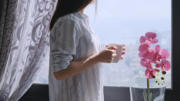 Millenial brunette woman holding a cup of tea or coffee and looking in the window alt
