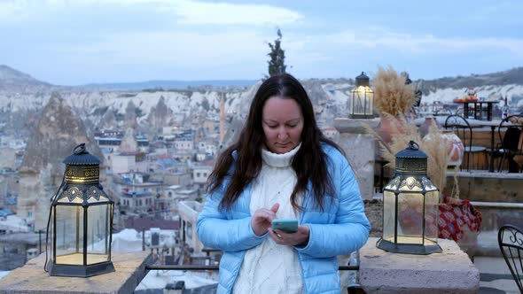 A Young Woman in a Blue Jacket Stands in the Evening and Looks at the Phone alt