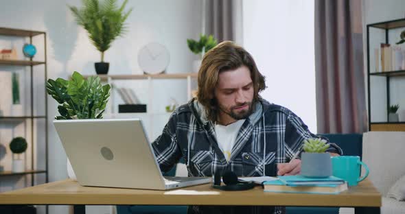 Man Sitting at the Desk at Home and Working with Computer and Papers alt