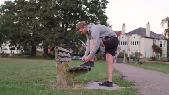 Young Attractive Man Tying His Shoes on a Bench Before He Sets of For a Run, In Slow Motion alt