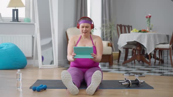 Excited Obese Woman Laughing Out Loud Sitting on Exercise Mat Watching Comedy Movie Online on Tablet alt