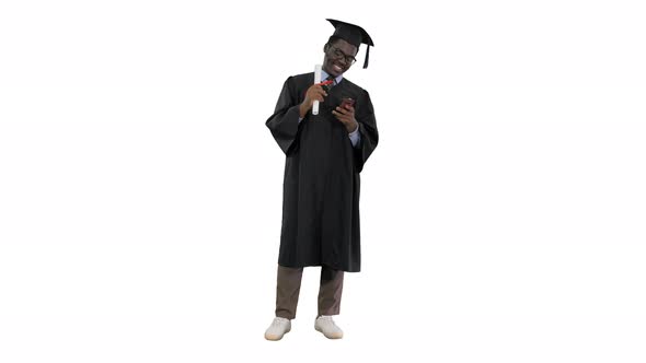 Smiling African American Male Student in Graduation Robe with Diploma Texting on the Phone on White
