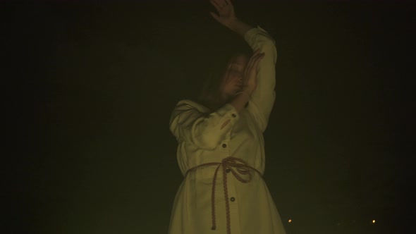 Slow ecstatic dance of young lady in white dress in darkness before fire yellow light low angle view alt