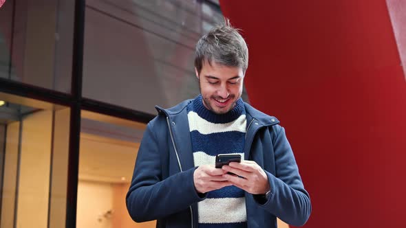 Handsome Young Man, Standing Outside the Office Building, Using Social Networks on Mobile Phone alt