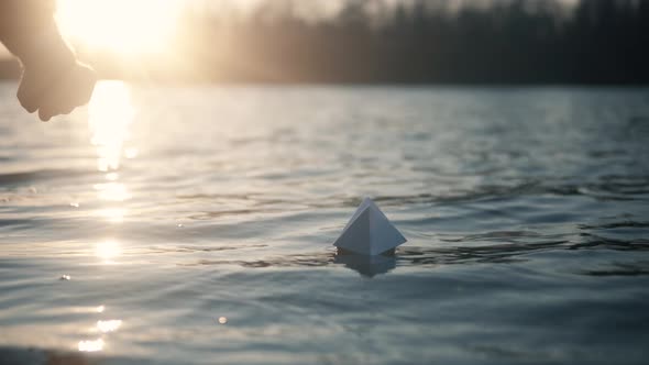 Paper Boat On River At Sunset. Origami Ship Sailing On Pond. Man Hand Launch Paper Boat On Water. alt
