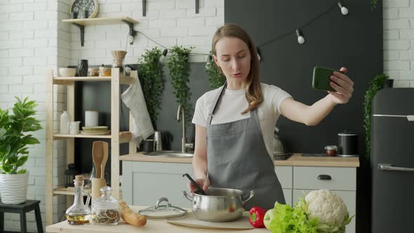 Happy Positive Female Blogger in an Apron Records a Cooking Video Blog on Her Mobile Phone alt