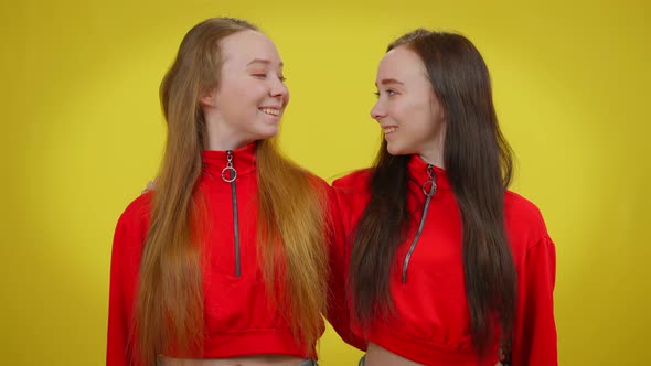 Cheerful Young Female Twins Looking at Each Other Turning to Camera Smiling Gesturing Forefinger Up alt