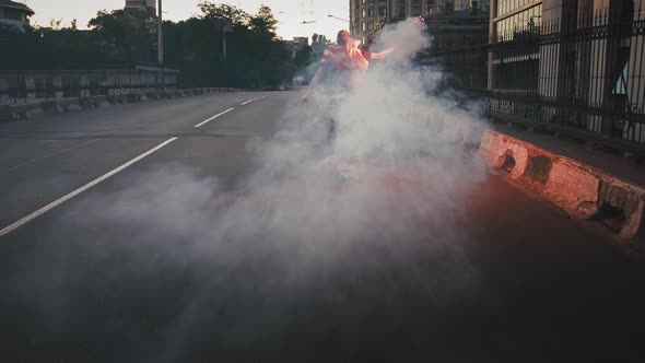 Young Guy in Flag of USA Tied on His Neck is Skateboarding Along Street with Burning Red Signal alt