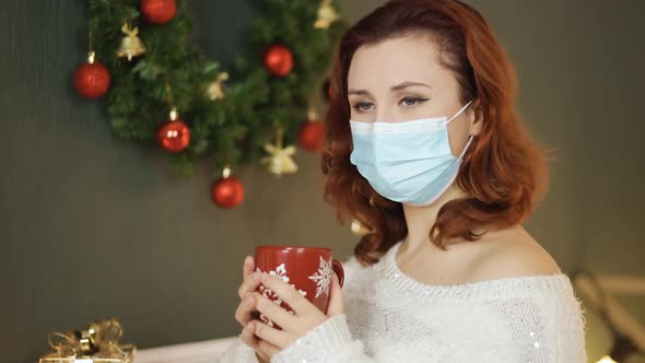 Sad Young Woman in Medical Mask Holds Red Christmas Cup of Tea alt