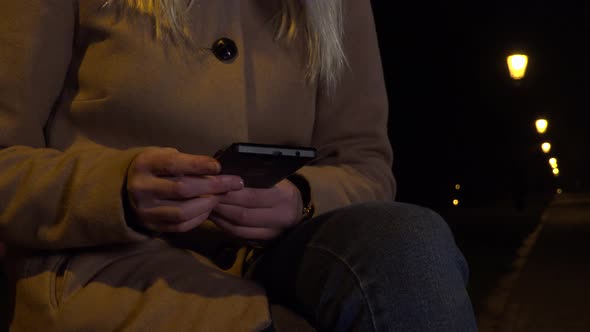 A Woman Sits on a Bench and Works on a Smartphone in an Urban Area at Night - Closeup alt