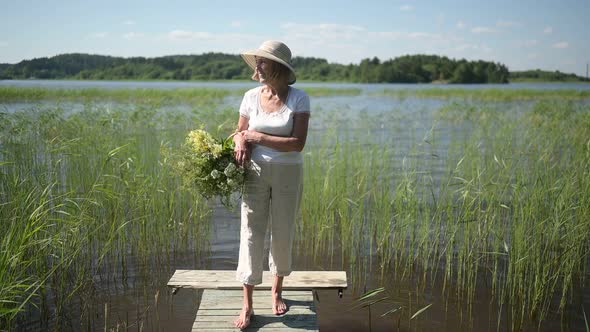 Happy Smiling Elderly Senior Woman in Straw Hat Walking Along Wooden Pier By Lake with Flowers in