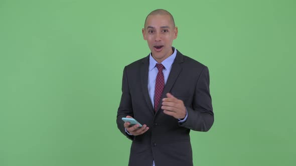 Happy Bald Multi Ethnic Businessman Using Phone and Looking Surprised alt