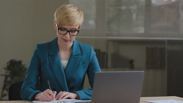 Middle Aged Female Entrepreneur Wears Glasses Using Laptop Writing Notes Sitting at Office Table alt