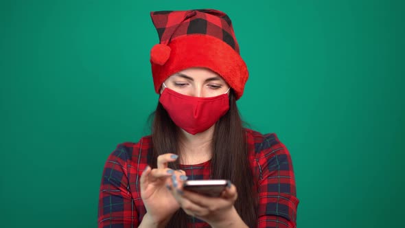 Woman in Santa Claus Hat, Wearing a Medical Mask on Her Face, Writes a Message on a Smartphone or
