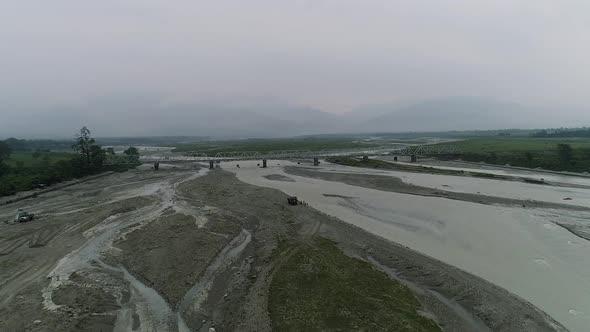 Aerial view of bridge over riverbed swollen by rushing water of rainy season, misty horizon of mount alt