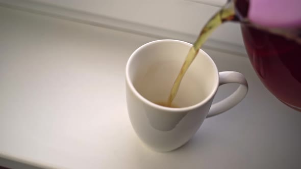 Human Pours Fragrant Tea From Glass Teapot Into White Ceramic Mug