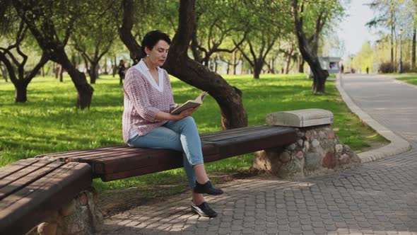 A Woman is Sitting on a Park Bench Reading a Book