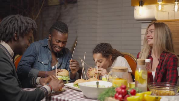 Happy Multiethnic People Eating Picnic Burgers Talking and Laughing Sitting at Evening Table alt