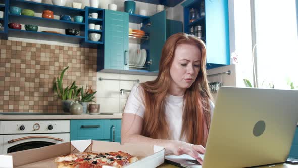 Focused woman working in the kitchen at the computer and snacking on pizza alt