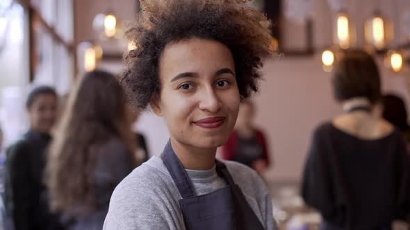 Close Up Footage of Dark Skinned Female with Afro Haircut Standing in Crowded Restaurant Smiling in alt