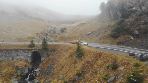 Aerial Shot of Orange Car Riding Through Mountain Road at Autumn alt