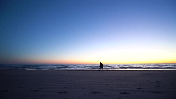 Traveler With Backpack Silhouette Walking On The Beach Near Sea After Sunset
