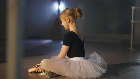 Side View Wide Shot of Happy Caucasian Girl in Tutu Smiling Admiring Golden Pointes Sitting on Floor alt