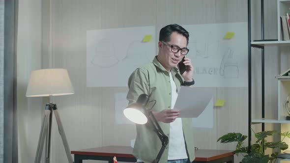 Asian Man Designer Talking On Smartphone While Looking At The Layout Bond In Hand And Walking alt