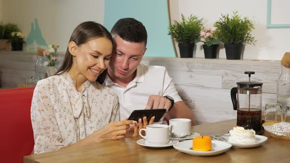 Girl and Guy Looks at Mobile Phone at Table in Sweet Shop alt