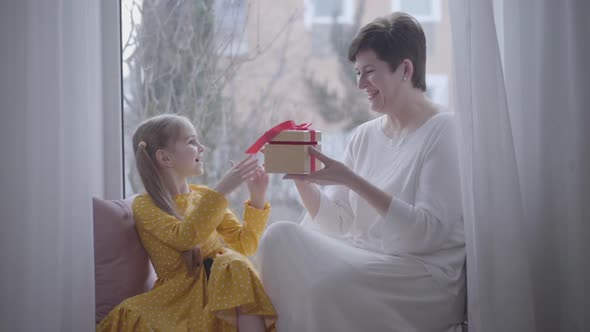 Cheerful Caucasian Girl Giving Gift To Positive Grandmother. Adult Brunette Woman and Pretty Little alt