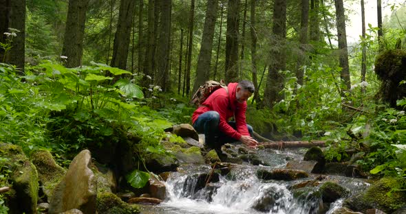 Bearded Man with a Backpack Washes His Hands with Water From a Mountain Stream alt