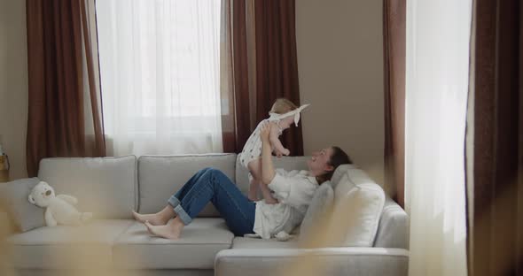 Mother Plays with Her Adorable Baby Daughter on a Sofa at Home alt