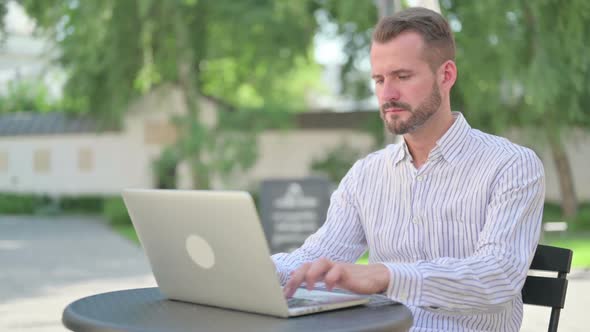 Middle Aged Man with Laptop Looking at Camera in Outdoor Cafe alt
