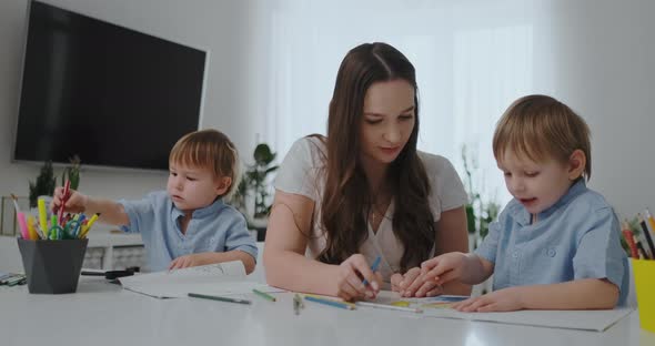 A Young Mother with Two Children Sitting at a White Table Draws Colored Pencils on Paper alt