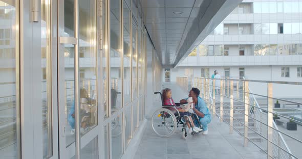 Laughing Little Girl Sitting on Wheelchair Playing with Nurse Outside Hospital alt