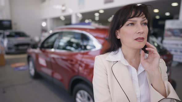 Thoughtful Caucasian Successful Woman Standing in Car Dealership Choosing New Vehicle alt