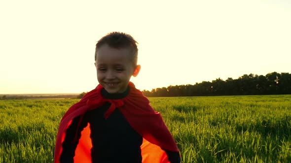 A Happy Child in a Superhero Costume in a Red Cloak Runs on Green Grass on a Sunset Background in alt