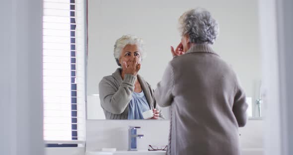 African american senior woman applying face cream while looking in the mirror at home alt
