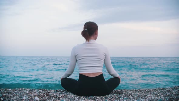 A Woman Sits on the Pebble Beach in the Lotus Position and Looks at the Sea alt