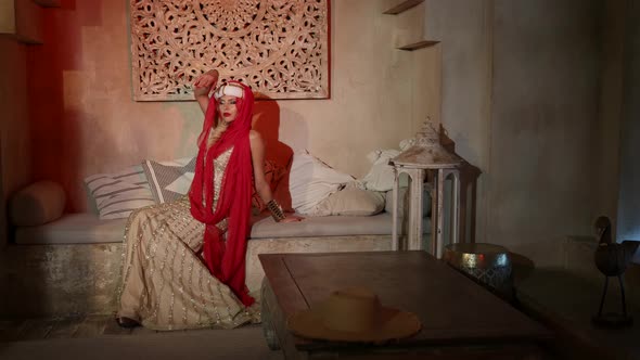 a Woman in a Turban a Red Scarf and a Sequined Dress is Sitting on a Sofa with Pillows in the Sunset alt