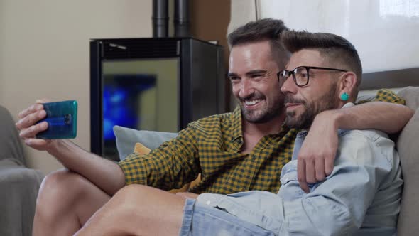Gay Couple Sitting in the Sofa At Home Making Video Call On Mobile Phone alt