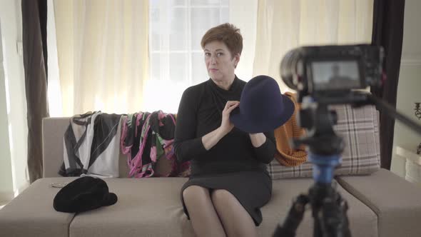 Portrait of Positive Senior Caucasian Woman Showing at Camera Hats and Scarfs. Mature Woman Sitting alt