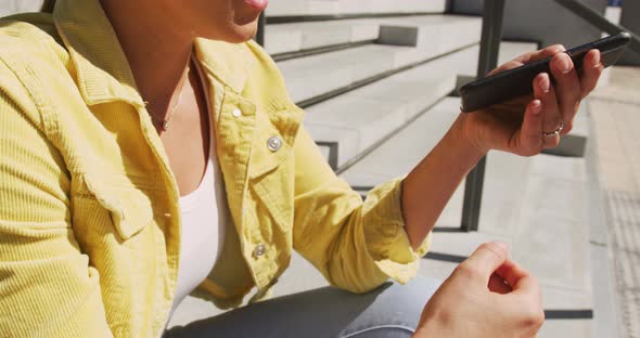 Happy caucasian woman sitting on stairs, using smartphone on sunny day alt