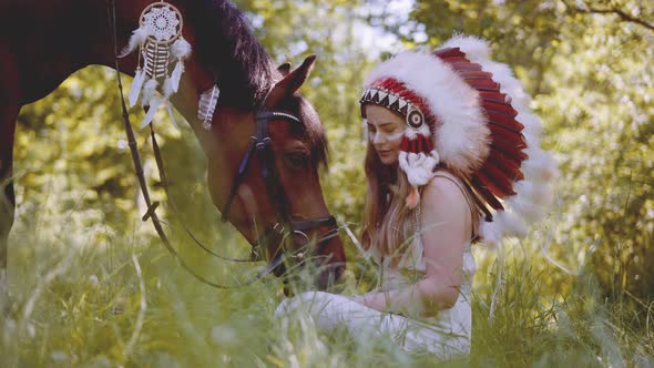Woman In Headdress Sitting And Feeding Horse In Sunlit Forest alt