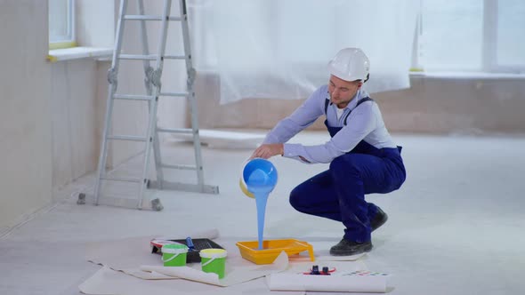 Man in Work Clothes and Cap Pours Blue Dyestuff in Paint Tray for Painting Walls During Renovation alt