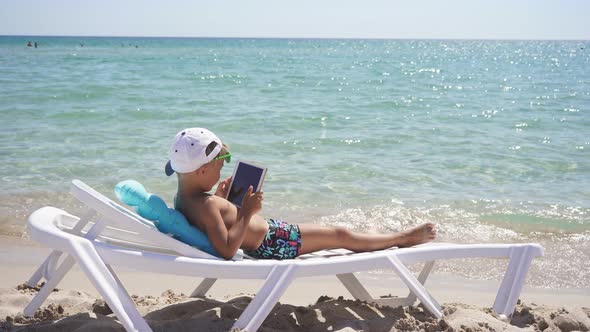Child is Relaxing on the Beach and Playing with a Tablet While Lying on a Chaise Longue