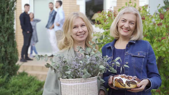 Two Caucasian Mature Women Smiling and Holding a Cake and a Bucket of Flowers alt